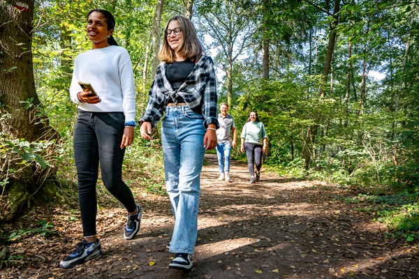 Twee jongeren wandelen ontspannen over een bospad omringd door groen in natuurgebied het Leudal met o0p de achtergrond de ouders.