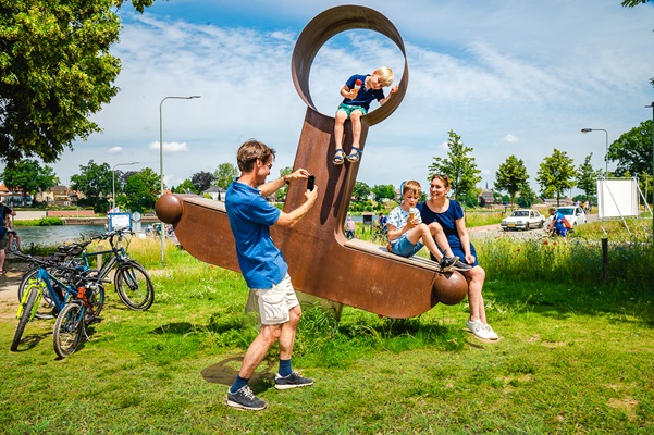 Een gezin maakt een foto bij een groot kunstwerk langs de Maasfietsroute, met fietsen geparkeerd in het gras.