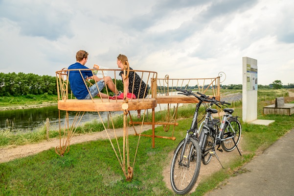 Twee mensen zitten op een kunstwerk met uitzicht over de Maas, naast een informatiepaneel.