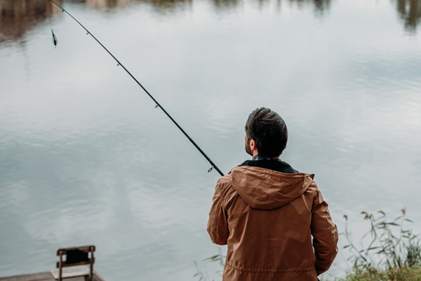 A man in a brown jacket is fishing at the edge of calm water.