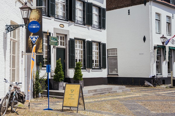Frontage of VVV Thorn, located in a white historic building with green shutters. In front of the entrance are an information board and a VVV flag. To the left of the door hangs a blue VVV sign and a banner of Museum Thorn. A bicycle leans against the wall on the cobbled street.