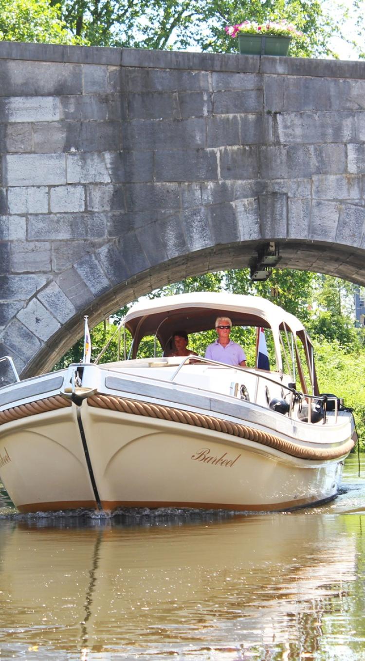Twee mensen varen met een beige sloep onder de stenen brug in Roermond, omgeven door groen en zonlicht.