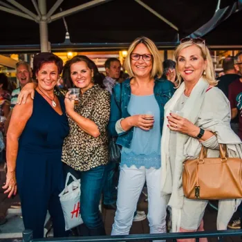 Vier lachende vrouwen met drankjes in de hand tijdens een gezellige avond uit op een terras.