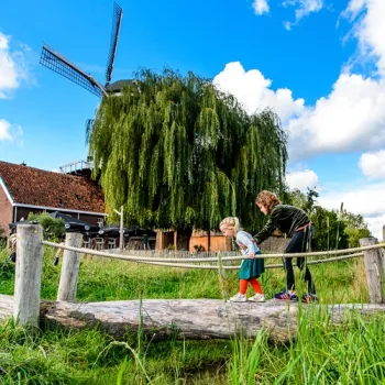 Erwachsener und Kind stehen auf einer kleinen Holzbrücke im Grünen, mit einer Windmühle im Hintergrund.